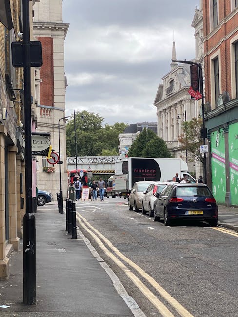 A narrow urban street scene near Shoreditch High Street station during daytime, showing several parked cars along the right side of the road and a large white moving truck positioned across the street