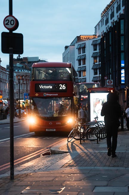 A narrow urban street scene near Shoreditch High Street station during daytime, showing several parked cars along the right side of the road and a large white moving truck positioned across the street