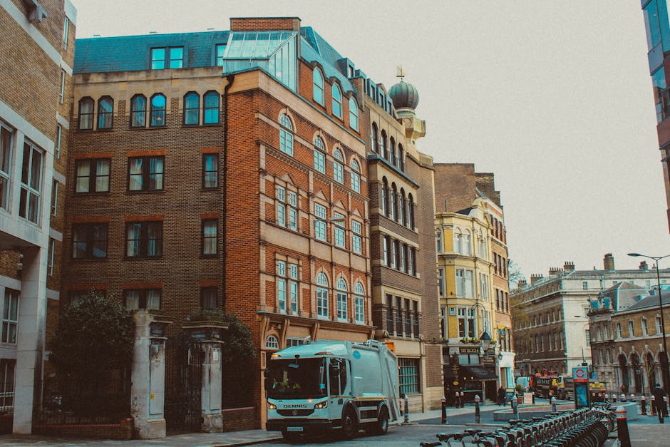 A large green delivery van with a closed cargo area is parked on a city street in Shoreditch, London, near several tall modern skyscrapers and older historic buildings. The van is positioned close to 